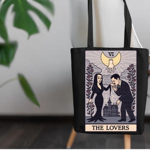 Addams Family tarot tote bag featuring Morticia & Gomez as The Lovers. Gothic artwork with roses, crescent moon, and haunted mansion. Perfect for tarot collectors and witchy Halloween fans.