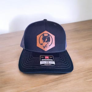 May include: Navy blue trucker hat with a brown leather patch featuring a cartoon wolf. The hat has a curved brim and a mesh back. The text "RICHARDSON" and "ORIGINAL 112 TRUCKER" are visible.