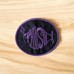 May include: Oval patch with a black background and a purple embroidered design. The design features the word "FISH" stylized as a fish, with bubbles. The patch has a purple border and is set against a wooden surface.