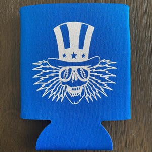 May include: Blue neoprene can cooler with a white graphic of a skull wearing a white top hat with stars and sunglasses. The skull has lightning bolt hair.
