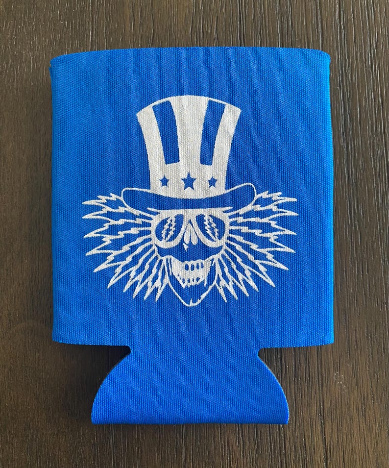 May include: Blue neoprene can cooler with a white graphic of a skull wearing a white top hat with stars and sunglasses. The skull has lightning bolt hair.