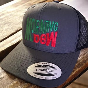 May include: A grey and black trucker hat with a red and green embroidered logo that says "Morning Dew". The hat has a black mesh back and a grey front with a silver sticker that says "The Authentic Yupoong Snapback Yupoong Since 1974".