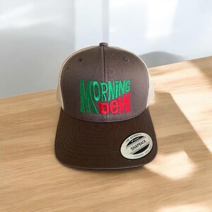 May include: A brown and tan baseball cap with a green and red embroidered logo that says "MORNING DEW". The cap has a mesh back and a snap closure. The label on the cap says "THE AUTHENTIC SNAPBACK YUPOONG SINCE 1974".