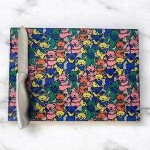 May include: A rectangular cutting board with a colorful pattern of dancing bears in various colors, including pink, yellow, blue, and green. A silver knife rests on the board. The board is on a white marble surface.