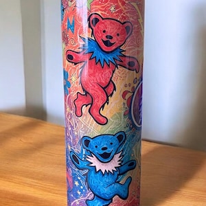 May include: A stainless steel tumbler featuring a colorful design with dancing bear illustrations. The bears are red and blue with white collars, set against a vibrant background of stars and psychedelic patterns. The tumbler is cylindrical.