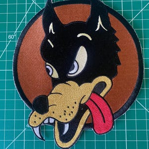 Op de afbeelding: Geborduurde patch met een cartoon wolfskop met een grimmige uitdrukking. De wolf is zwart met een bruine snuit en een rode tong. Het is geplaatst tegen een bruine cirkelvormige achtergrond met een zwarte rand. De patch ligt op een groene snijmat.