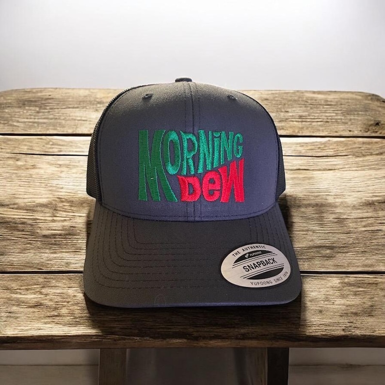 May include: A gray and black trucker hat with a green and red embroidered logo that says "Morning Dew". The hat has a black mesh back and a gray snap closure.