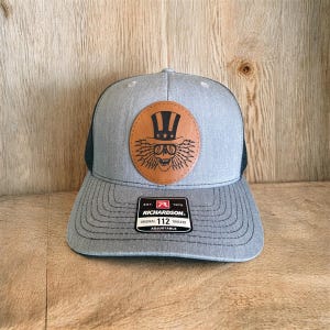 Pode incluir: Boné cinza de caminhoneiro com parte traseira em tela preta e aba curva. O boné apresenta um patch redondo de couro marrom com uma caveira usando uma cartola. O patch é centralizado na frente. O boné tem o texto "RICHARDSON" e "ORIGINAL 112 TRUCKER".