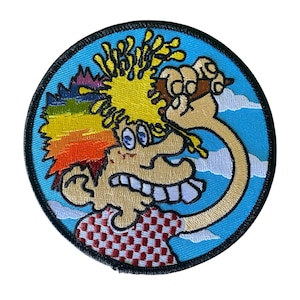 May include: Circular embroidered patch with a cartoon character. The character has yellow hair, a rainbow section, and a surprised expression. It wears a red and white checkered shirt. The patch has a blue background with white clouds and a black border.
