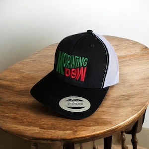 May include: Black and white trucker hat with a green and red embroidered logo that says "MORNING DEW". The hat has a black brim and a white mesh back.