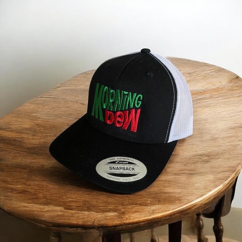 May include: Black and white trucker hat with a green and red embroidered logo that says "MORNING DEW". The hat has a black brim and a white mesh back.