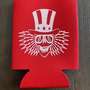 May include: Red neoprene can cooler with a white graphic of a skull wearing a white Uncle Sam hat and sunglasses. The skull has a white lightning bolt design around it.