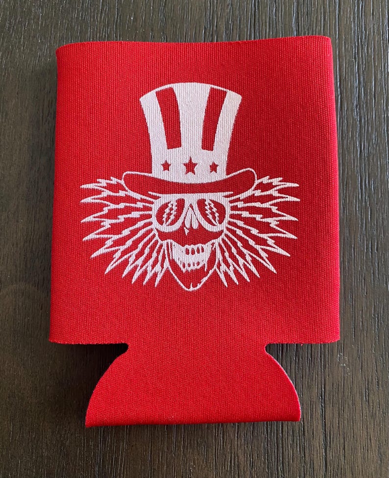 May include: Red neoprene can cooler with a white graphic of a skull wearing a white Uncle Sam hat and sunglasses. The skull has a white lightning bolt design around it.