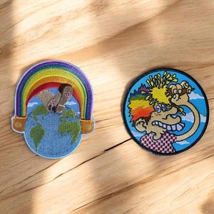 May include: Two embroidered patches on a wooden surface. One patch features a rainbow arching over a globe, with a figure on top. The other patch is circular, depicting a cartoon character with wild hair and a checkered shirt.