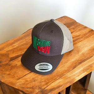 May include: A brown and beige trucker hat with a green and red embroidered logo that says "MORNING DEW". The hat has a black adjustable strap with a label that says "THE AUTHENTIC SNAPBACK YUPOONG SINCE 1974".