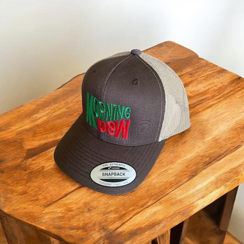 May include: A brown and beige trucker hat with a green and red embroidered logo that says "MORNING DEW". The hat has a black adjustable strap with a label that says "THE AUTHENTIC SNAPBACK YUPOONG SINCE 1974".