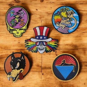 May include: A selection of embroidered patches with diverse designs. These include a skull with a rainbow design, a cartoon character with rainbow hair, a wolf, a cat, and a skull with dancing bears.