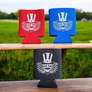 May include: Three different colored can coolers with a white graphic of a skull wearing a top hat with stars and stripes. The skull has lightning bolt hair and is wearing sunglasses.