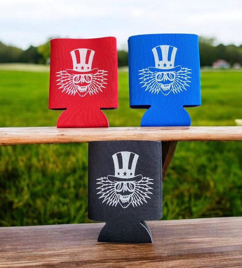 May include: Three different colored can coolers with a white graphic of a skull wearing a top hat with stars and stripes. The skull has lightning bolt hair and is wearing sunglasses.