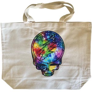 May include: Cream-colored canvas tote bag featuring a colorful skull design. The skull is filled with a galaxy pattern in shades of blue, green, yellow, and purple, with a black lightning bolt. The bag has beige handles.