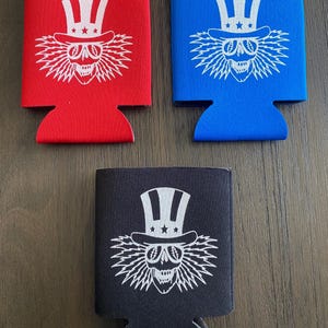 May include: Three different colored can coolers with a white skull wearing a red, white, and blue top hat with stars. The skull has lightning bolt hair and is smiling. The can coolers are red, blue, and black.