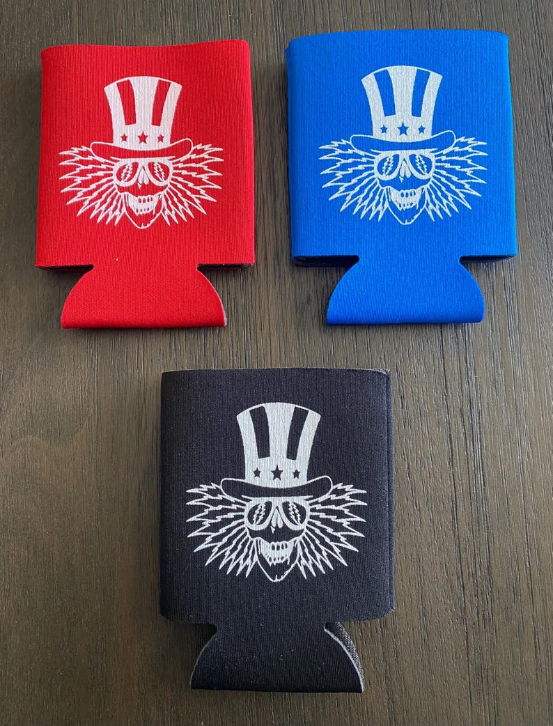 May include: Three different colored can coolers with a white skull wearing a red, white, and blue top hat with stars. The skull has lightning bolt hair and is smiling. The can coolers are red, blue, and black.