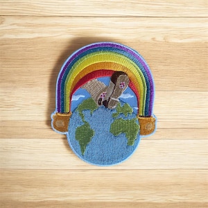 May include: Embroidered patch featuring a rainbow arching over a blue globe. The rainbow displays vibrant colors, with a pot of gold at each end. Two socks are positioned on top of the globe. The patch is on a wooden surface.
