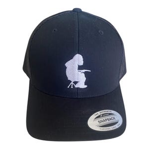 Peut inclure: Casquette de camionneur noire avec une silhouette blanche brodée d'une personne assise sur un tabouret, tenant une canne à pêche. La casquette a une visière incurvée et un dos en filet. Fermeture Snapback.