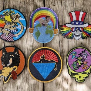 May include: Six embroidered patches with diverse designs. The patches depict cartoon characters, a rainbow over the Earth, a skull in a patriotic hat, a wolf, a cat, and dancing bears. The patches are circular and have a stitched border.