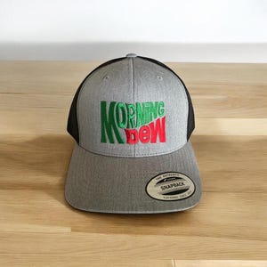 May include: A grey and black baseball cap with a green and red embroidered logo that says "MORNING DEW". The cap has a black mesh back and a grey front with a black brim. The cap has a small white tag on the back that says "THE AUTHENTIC SNAPBACK YUPOONG SINCE 1974".