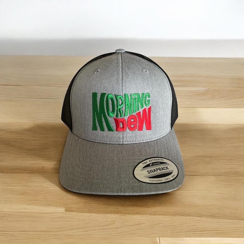 May include: A grey and black baseball cap with a green and red embroidered logo that says "MORNING DEW". The cap has a black mesh back and a grey front with a black brim. The cap has a small white tag on the back that says "THE AUTHENTIC SNAPBACK YUPOONG SINCE 1974".