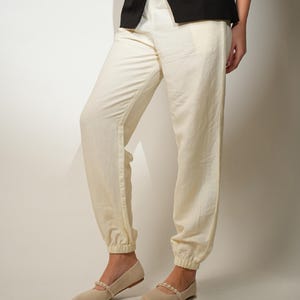 May include: Cream-coloured, loose-fitting trousers with elasticated cuffs and a matching top. The trousers are paired with beige flats adorned with a pearl strap. A black waistcoat is worn over the top. The outfit is suitable for casual wear.