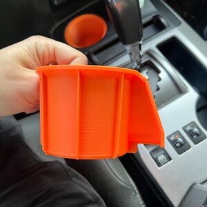May include: Orange 3D printed cup holder insert for a car. The insert has a lip on one side to help keep drinks from spilling.