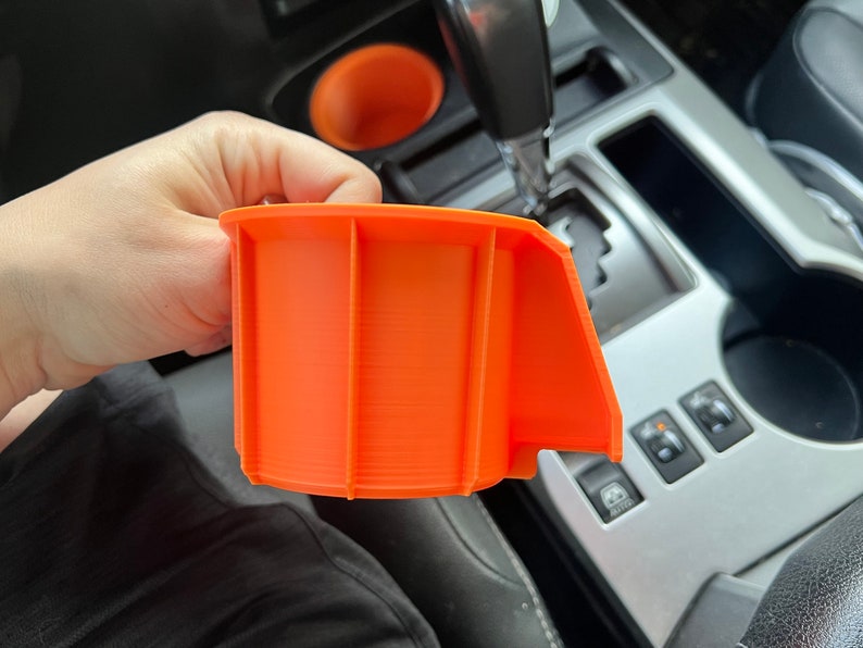 May include: Orange 3D printed cup holder insert for a car. The insert has a lip on one side to help keep drinks from spilling.