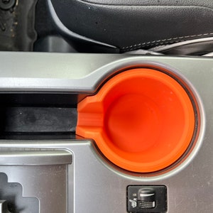 May include: A close-up of a car's center console with a gray and black interior. The console features a cup holder with an orange silicone insert.