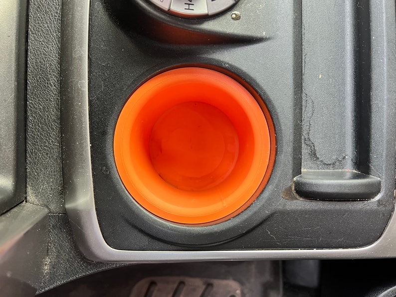 May include: A close-up of an orange cup holder in a car. The cup holder is made of a soft, flexible material and is designed to fit standard-sized cups.