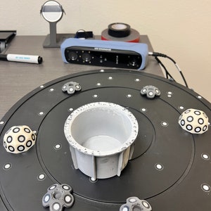 May include: A black turntable with a white circular pattern and a gray, cylindrical object in the center. There are six gray, circular objects with white circles around the edge of the turntable.