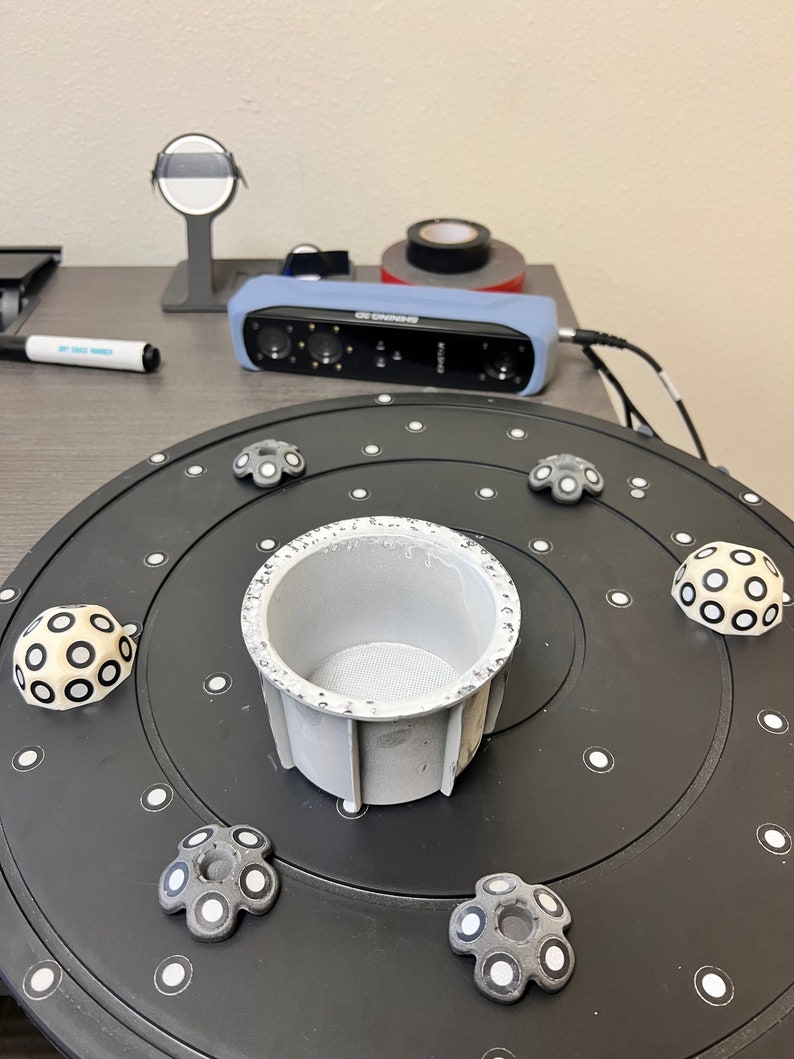 May include: A black turntable with a white circular pattern and a gray, cylindrical object in the center. There are six gray, circular objects with white circles around the edge of the turntable.