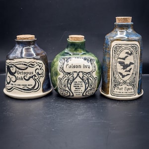 Könnte beinhalten: Drei Keramik-Trankflaschen mit Korken. Die Flaschen sind mit "Octopus Ink", "Poison Ivy" und "Bat Eyes" beschriftet. Die Flaschen haben ein rustikales, handgefertigtes Aussehen mit dunkelblauen, grünen und braunen Glasuren.