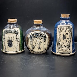 Puede incluir: Tres botellas de cerámica de boticario con tapones de corcho. Las botellas están etiquetadas como "Dried Beetle Arsenic", "Cobweb Oil" y "Genuine Witches Warts". Cada botella tiene una forma y un esquema de color únicos, con etiquetas en blanco y negro.