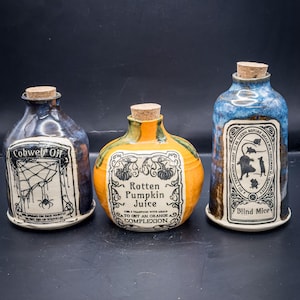 Könnte beinhalten: Drei Keramik-Trankflaschen mit Korken. Die Flaschen sind blau, orange und braun, jede mit einem einzigartigen Etikett mit Text und Illustrationen. Die Etiketten lauten "Cobweb Off", "Rotten Pumpkin Juice" und "Blind Mice".