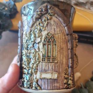 Könnte beinhalten: Handgefertigte Keramiktasse mit detailliertem, strukturiertem Design, das einer verwitterten Holztür mit gotischem Fenster ähnelt. Die Tasse hat eine braune und beige Farbpalette mit grünen Akzenten von Kletterpflanzen.