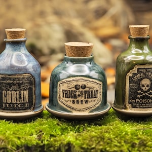 Puede incluir: Tres botellas de cerámica para pociones con tapones de corcho. Las botellas son azules, verdes y verde oliva, cada una con una etiqueta. Las etiquetas dicen "Goblin Juice", "Trick or Treat Brew" y "Poison". Las botellas están sobre un lecho de musgo verde.