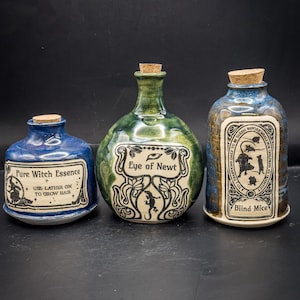 Puede incluir: Tres botellas de cerámica de pociones con tapones de corcho. Las botellas son azules, verdes y marrones con etiquetas que dicen "Pure Witch Essence", "Eye of Newt" y "Blind Mice". Las etiquetas presentan ilustraciones y texto.