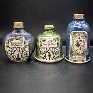 Puede incluir: Tres botellas de cerámica para pociones con tapones de corcho. Las botellas son azules, verdes y azules con etiquetas que dicen "Black Cat Whiskers", "Eye of Newt" y "Genuine Witches Warts". Las etiquetas tienen bordes decorativos e ilustraciones.