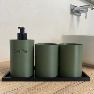 Könnte beinhalten: Ein Set aus drei grünen zylindrischen Badezimmerbehältern mit einem schwarzen Tablett. Der mittlere Behälter hat einen schwarzen Pumpspender und das Wort "Seife" auf der Vorderseite.
