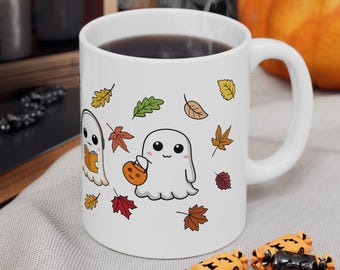 May include: White ceramic mug featuring a Halloween-themed design. The mug showcases two cartoon ghosts, one holding a pumpkin, surrounded by colorful autumn leaves. The mug is filled with dark liquid.