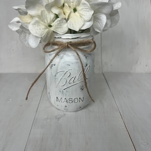 Könnte beinhalten: Ein weiß bemaltes Einmachglas mit einer braunen Kordelschleife und weißen Blumen. Das Glas trägt den Schriftzug "Ball Mason" auf der Vorderseite.