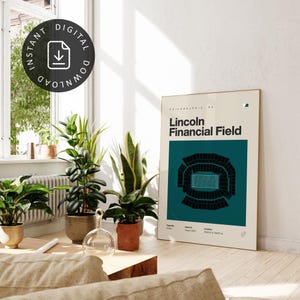 May include: A framed poster featuring the text "Lincoln Financial Field" with a teal and black stadium illustration. The poster is propped against a white wall, surrounded by potted plants and a window.