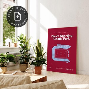 May include: A burgundy poster featuring a light blue stadium layout with the text "Dick's Sporting Goods Park" in white. The poster includes the location and year 2007. The image is in a well-lit room with plants.